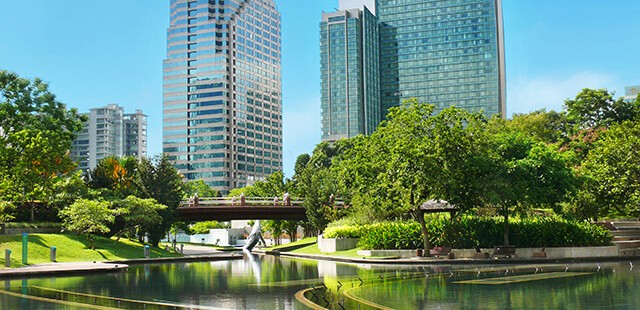 A view of two prominent office buildings surrounded by an urban park lush with greenery and a small pond. Park benches lines the walk ways surrounding the pond.