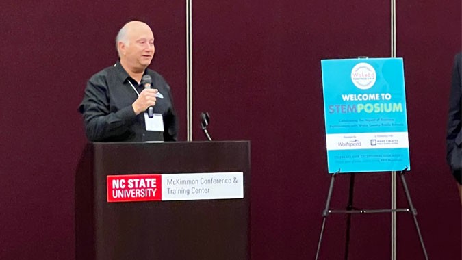 John Palmour holding a mic and standing behind a podium in a conference hall with a "Welcome to STEMposium" sign on his right.